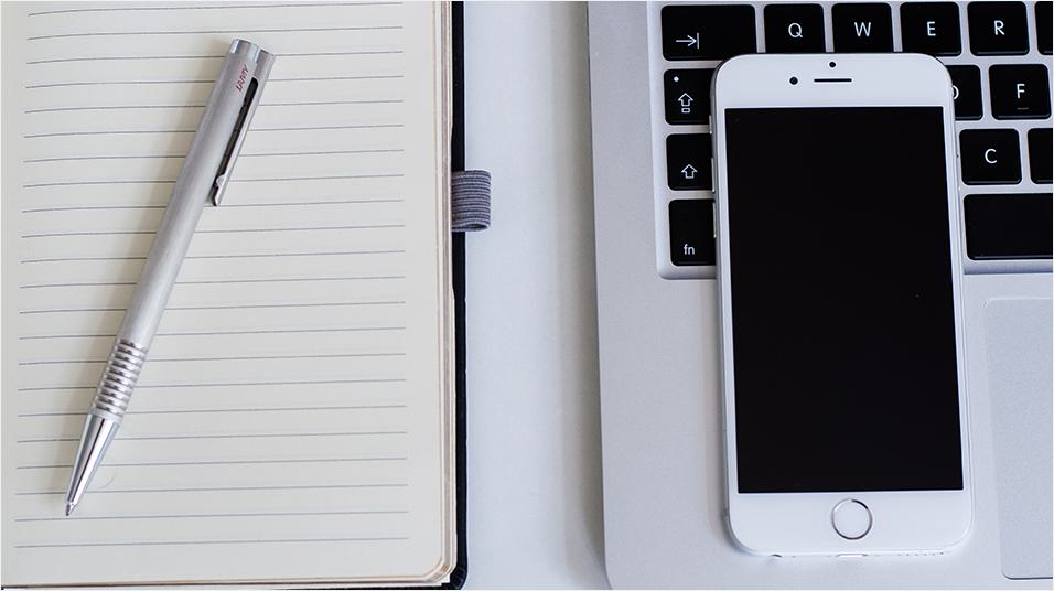 This is an image of a white lined notepad with a silver pen on top. On the right is a Macbook with just the keys visible, and a white iPhone on top.