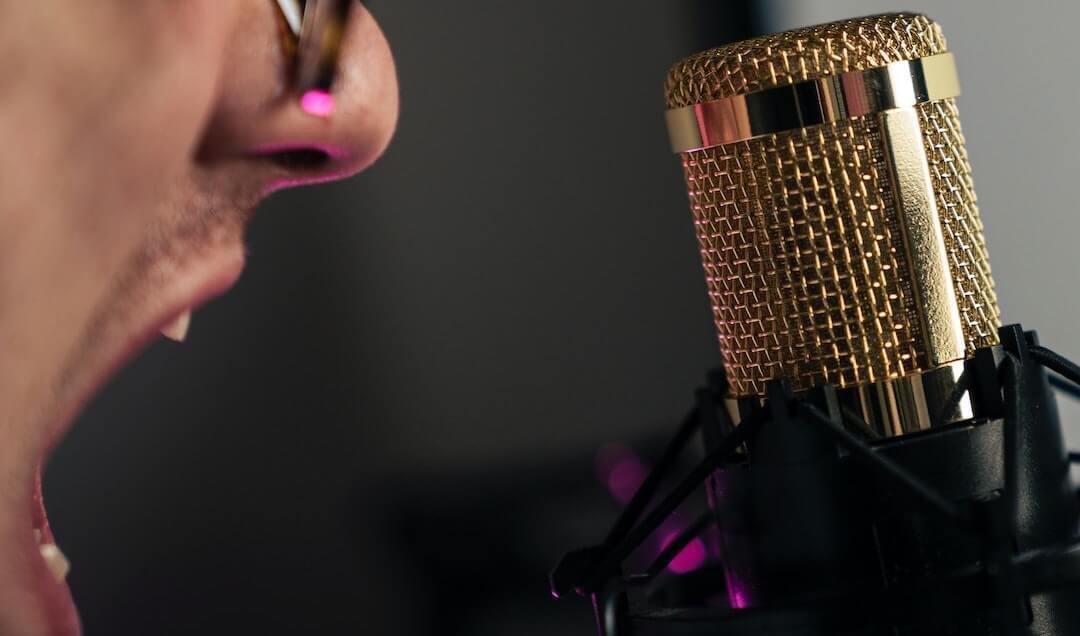 A white man wearing glasses opens his mouth in front of a microphone, appearing to be yelling. The microphone is gold.