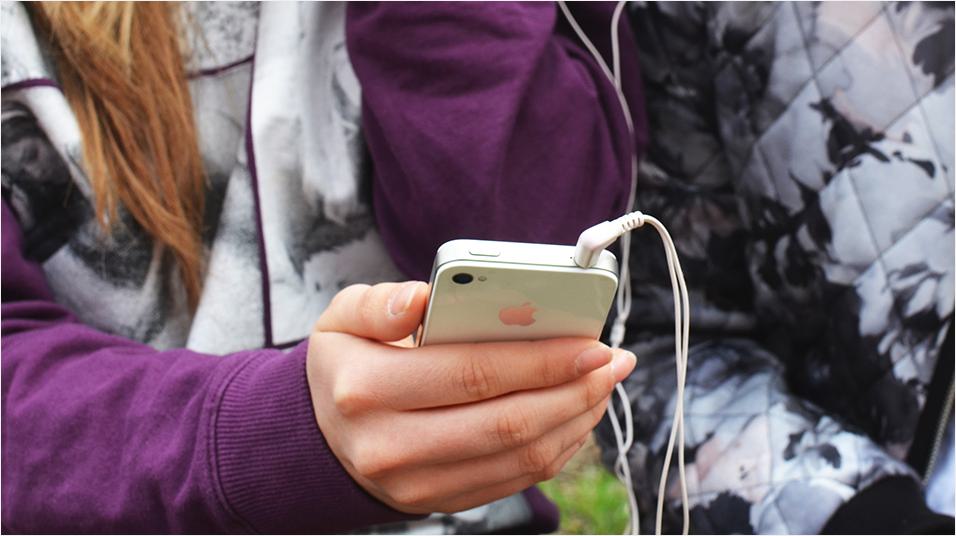 This is an image of a white hand holding an iphone. The iPhone has white headphones plugged into it. The person is wearing a purple hoodie, and has long sandy coloured hair. We can only see the shoulders and torso of the person.