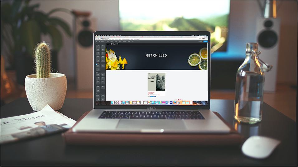 This is a picture of a Macbook on a table. On the left is a cactus in a white pot. On the right is an open glass bottle of water. In the background is a mic on a stand.