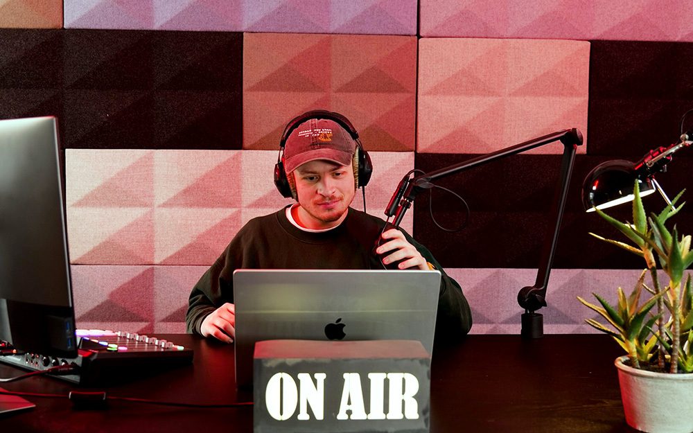 Image shows a man in a radio studio with a laptop, microphone, and mixing console.