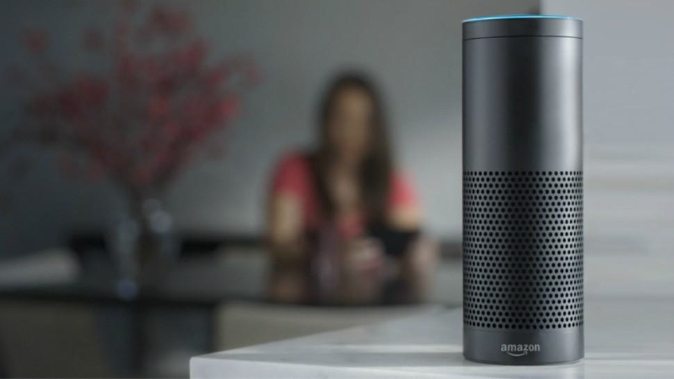 Image shows an Alexa-enabled smart speaker, on a desk. In the blurred background is a woman sat down and on her phone.