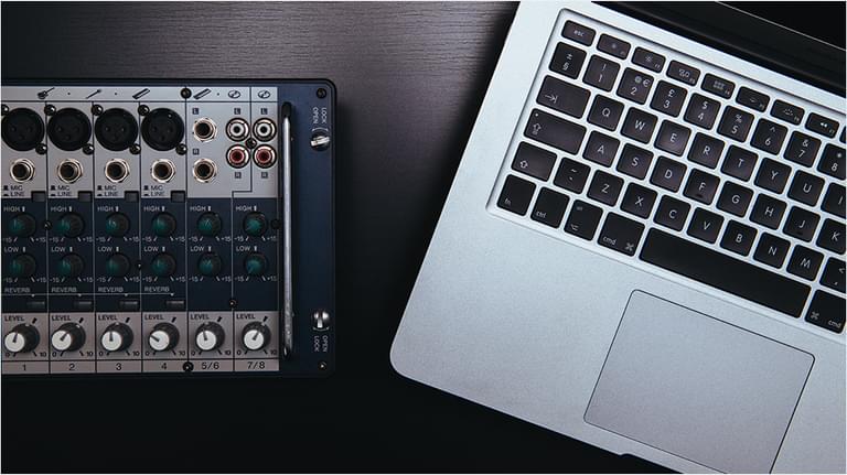 This is a mahogany dark desk. On the left is a mixing desk in black. On the right is a Silver and black Macbook keyboard and mouse set.