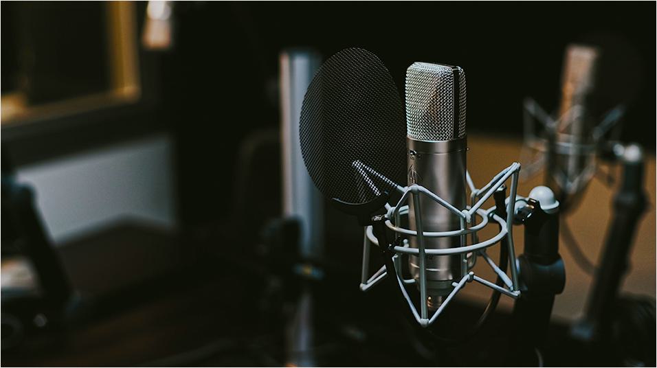 There is a black backdrop of a radio studio. There is a black mic on a stand visible in the centre of the image, with triangular elements visible.