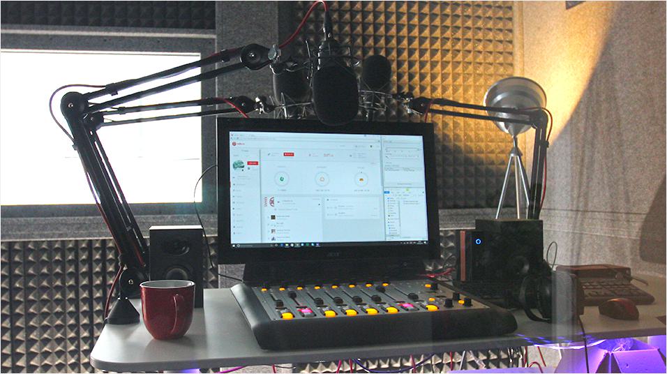 This is an image of a small home studio. In the background is warm light and soundpanels. In the foreground of the image is a monitor with a mixing desk in front of it.