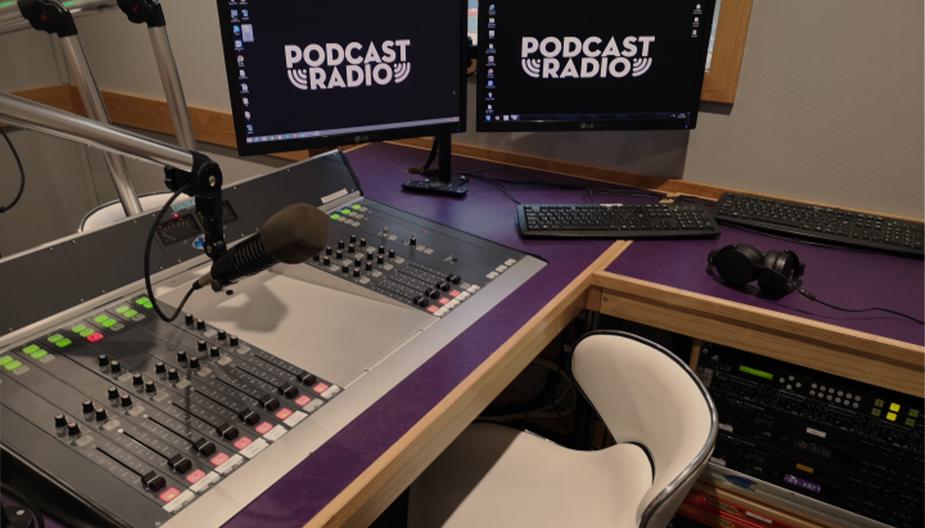 Image shows the inside of the Podcast Radio studio, with 2 broadcasting desks, 2 computer screens and a microphone on a desk.