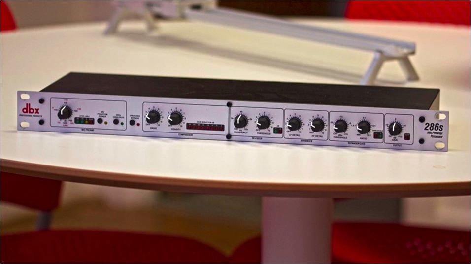 This is an image of the Dbx 286 S Microphone Processor, side on. It is a slim grey panel with various black knobs on it, on a white desk.