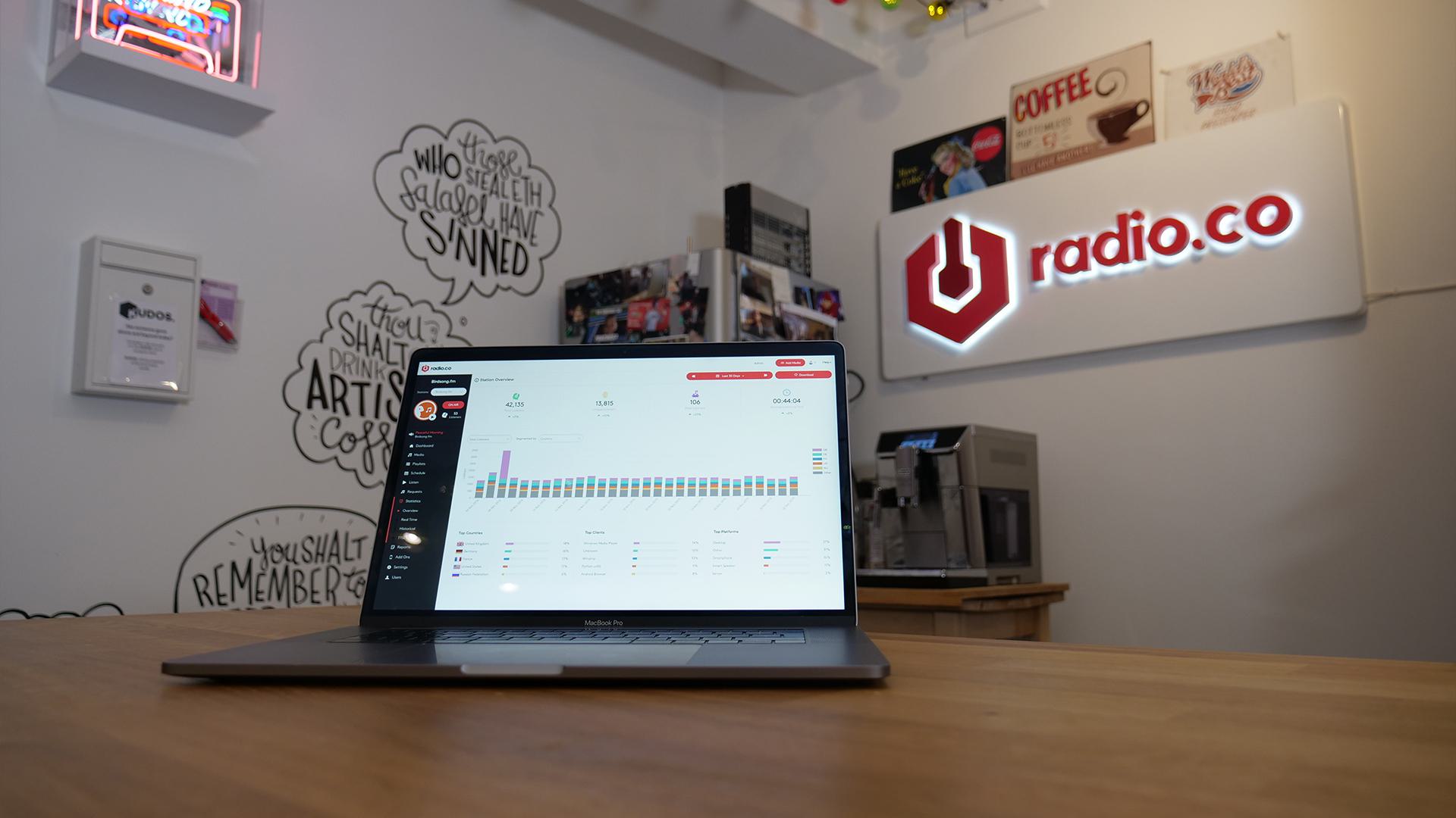 This is a white studio background with various signs and framed pictures on the wall, with a Macbook in the forefront of the image. The radio.co logo is on the wall in neon on the far right of the image.