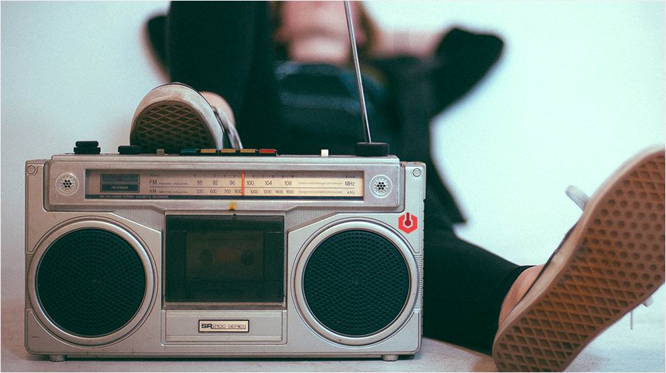 This is a neutral background with a person wearing sneakers and a black hoodie. They are sitting back listening to a silver boombox that's sat in front of them on the left.