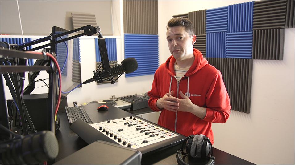 This is a picture of a white man with dark short hair wearing a red hoodie. He is looking into the camera. His hands are joined together over his chest. He stands in front of a mixing desk and a black microphone on a boom arm. The background is neutral.