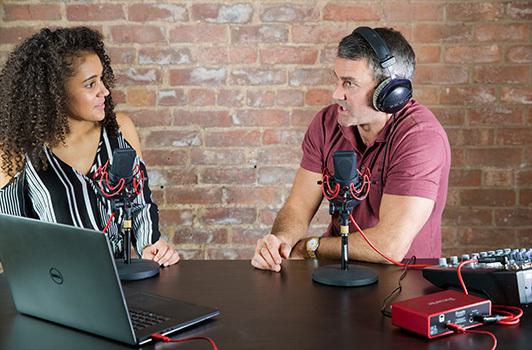 This is an image of a woman with long brown curly hair wearing a stripy top who sits in front of a microphone and a Macbook. She looks at her cohost, a man with grey hair and black headphones on. Both parties speak into microphones, conducting an interview or podcast. The background is a brick wall, plain.