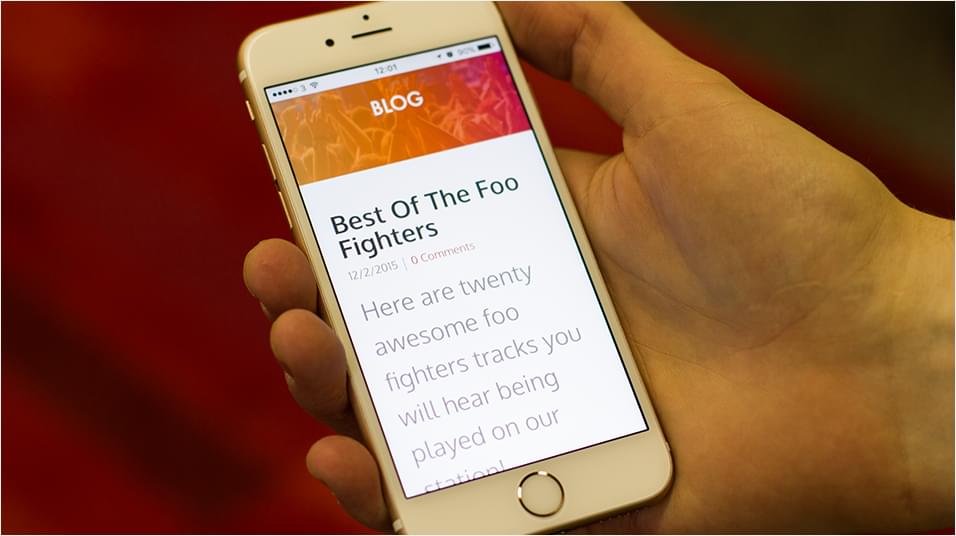 This is an image of a white iPhone in a hand against a red neutral backdrop. The phone has an orange banner and reads "BLOG' and below the blog topic is 'Best of the Foo Fighters' with a stock example of a blog article.