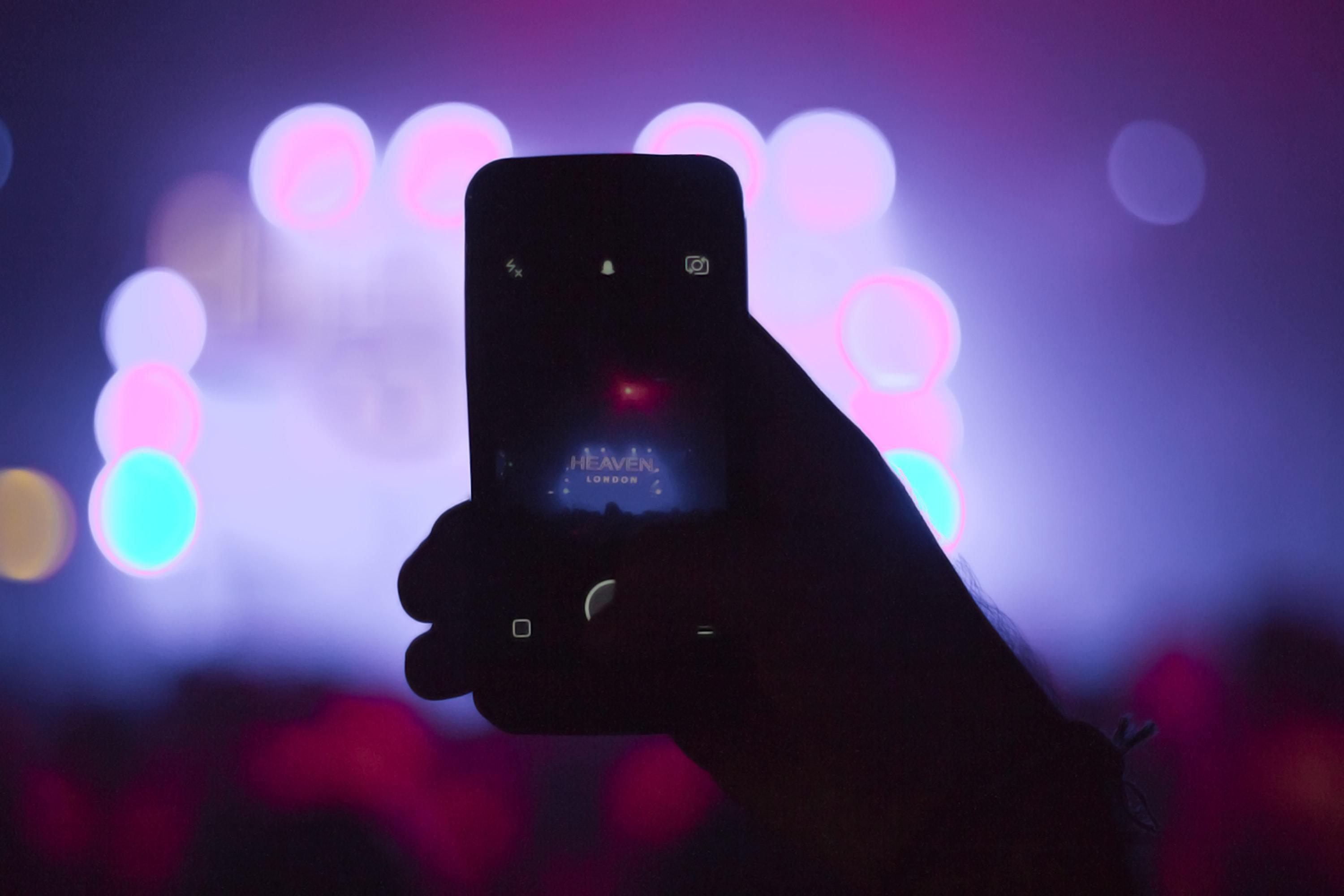 Image shows a hand holding up a smartphone and recording a night-time concert.