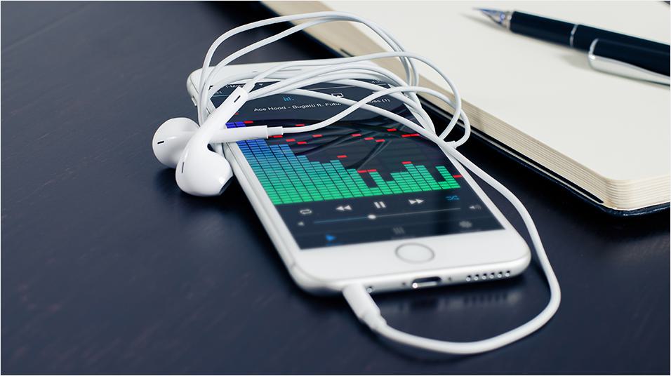 This is a black neutral backdrop with the corner of a Macbook visible. There is a white iphone in the middle with classic wired headphones connected and wrapped over the top. The screen reveals a green graph of sound levels in the middle.