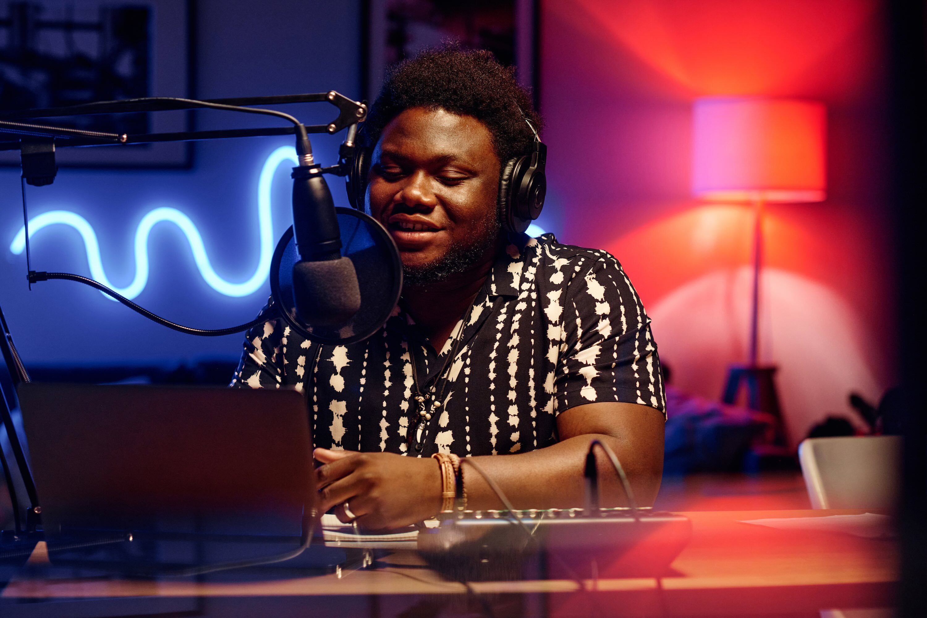 Male radio host wearing headphones speaks into a microphone in a studio, recording a live show on a laptop with professional audio equipment