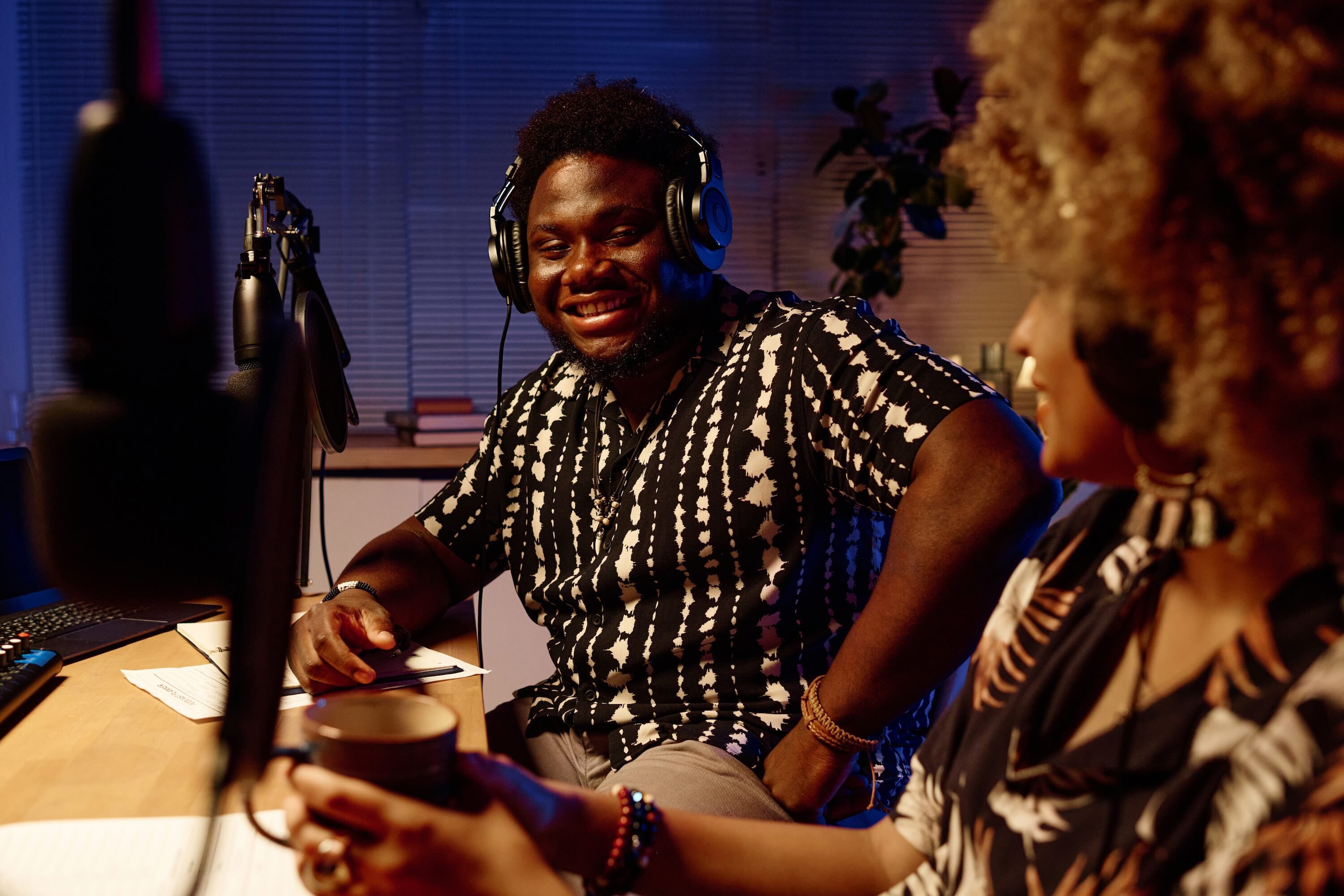 Radio host wearing headphones smiles while speaking with a female guest during an in-studio interview, microphones set up for a recorded conversation.