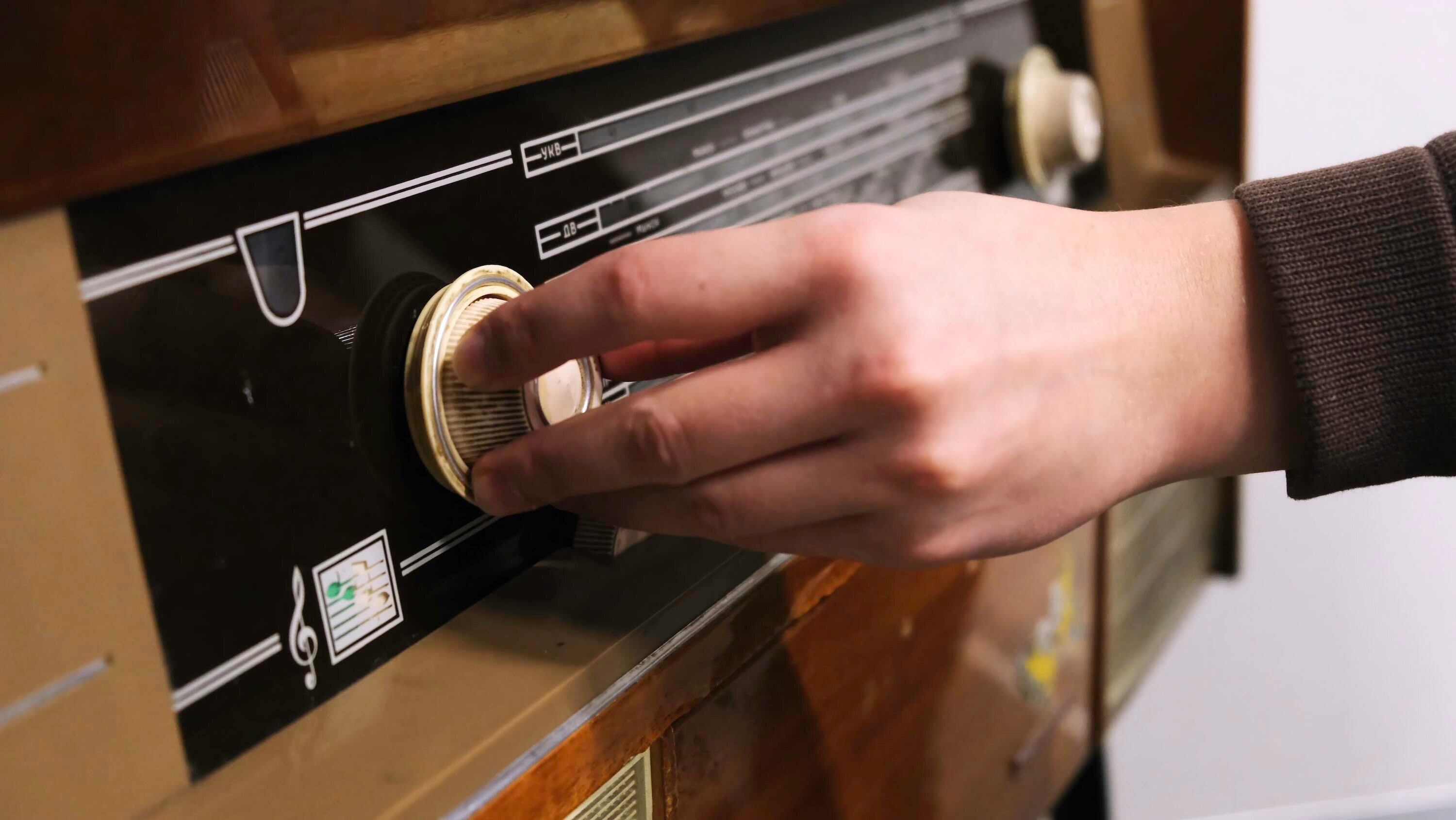 Close-up of a person’s hand turning the tuning knob on a vintage radio, with the frequency scale and musical note icon visible on the front panel.