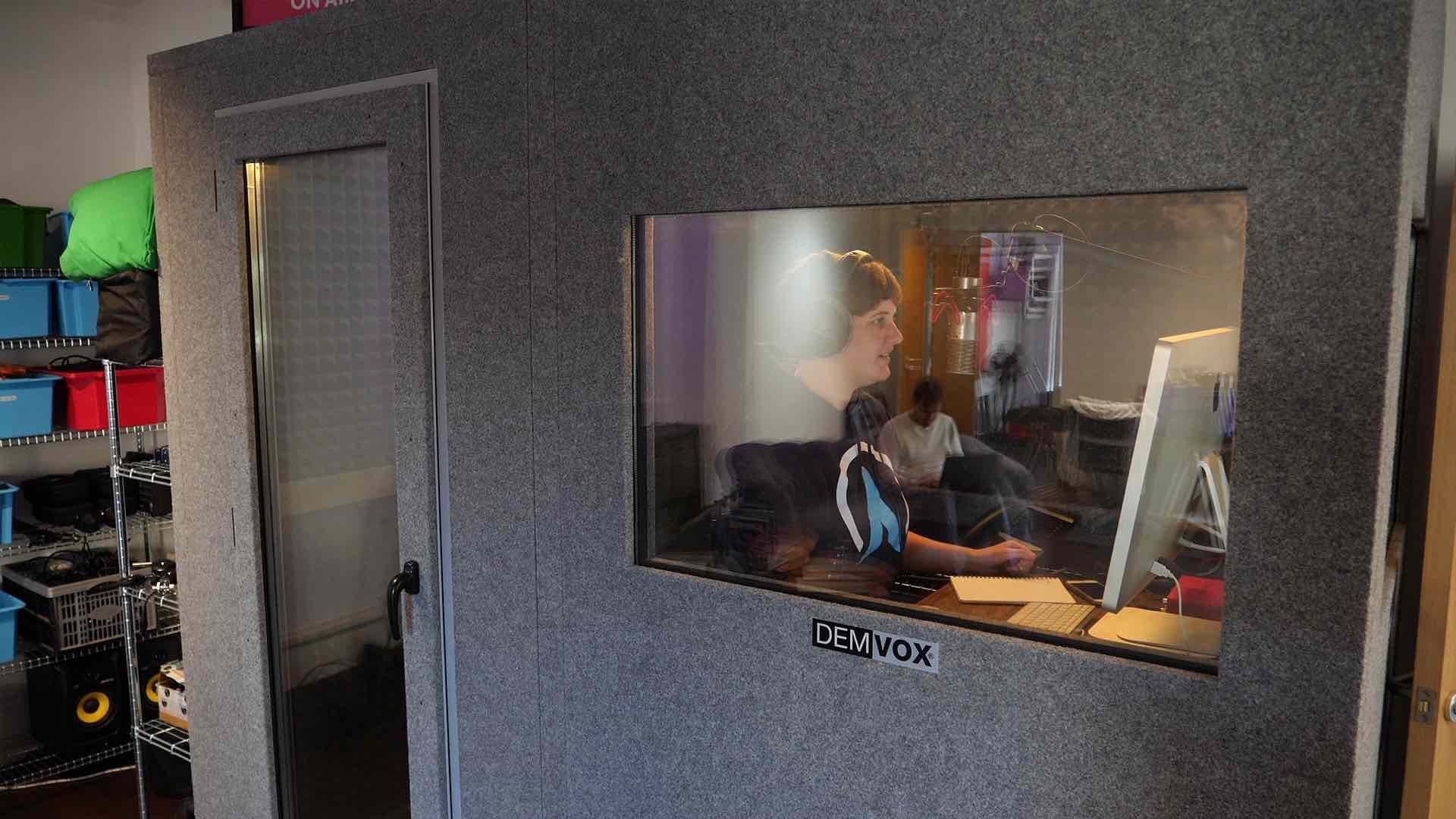 Image shows a stand alone radio booth, with one door and one window and a presenter in the booth speaking in a microphone.