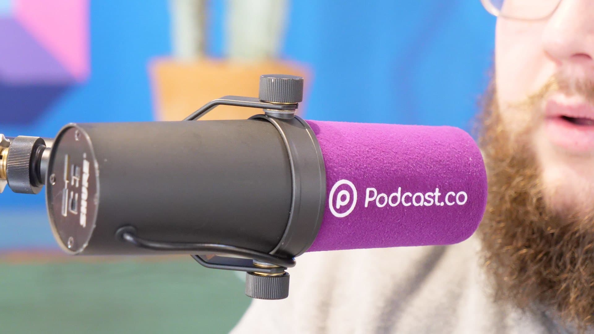 This is an image of a man with a ginger beard speaking into a purple and black Podcast.co microphone. The background is blue and purple and yellow, and is blurred out. We only see his mouth and the mic.