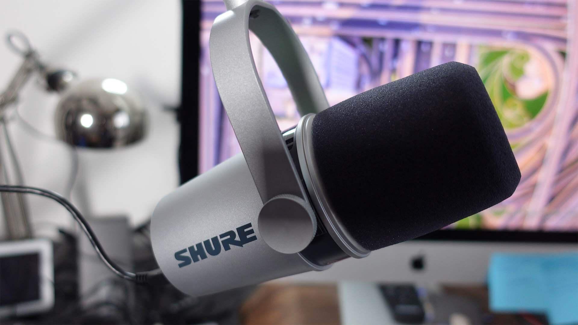 Image shows a silver Shure MV7 microphone, hanging from a mic stand and with a computer in the background.