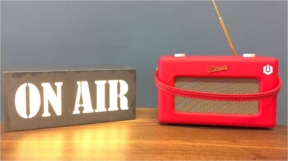 This is an iamge of a red Roberts radio on the right. On the left is a white neon ON AIR sign. The background is grey and neutral.