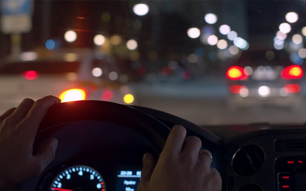Image shows a pair of hands on a steering wheel, driving in the dark.