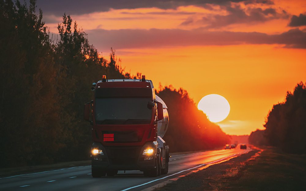 Image shows a truck driving down a highway with a large sunset in the background.