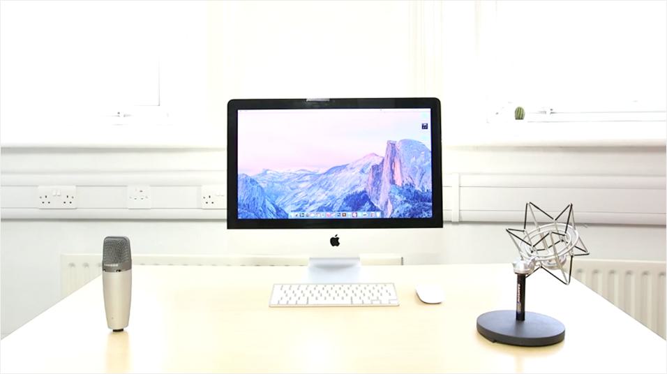 This is an image with a white backdrop with a silver Mac in the centre. The Mac's screen is a generic purple background. On the left is a silver Mic, on the black is an angular black lamp.