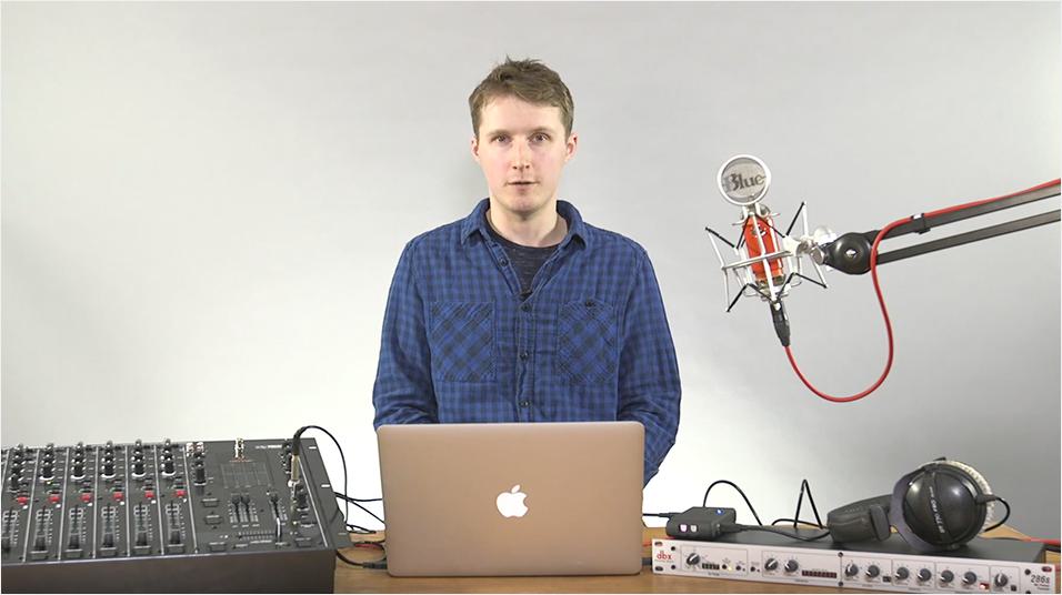 This is an image of a white man with short sandy blonde hair, looking into the camera and wearing a blue shirt. He is stood in front of a Macbook and speaking into a microphone on an arm. On the left is a black mixing desk on the right is a keyboard.