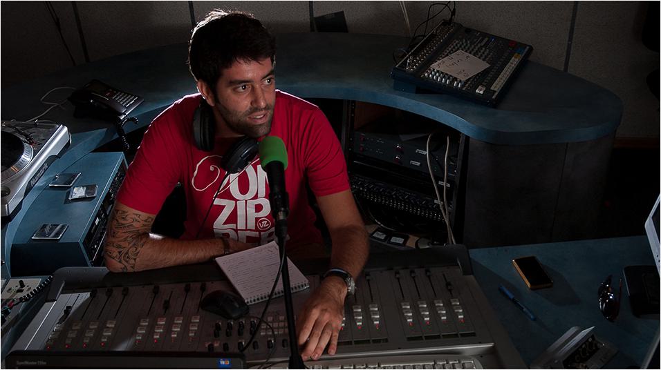 This is an image of a man with black hair and a black beard, sat in front of a mixing desk and a green microphone. He is presenting radio. The background is a blue curved desk typiacl of a studio setup.