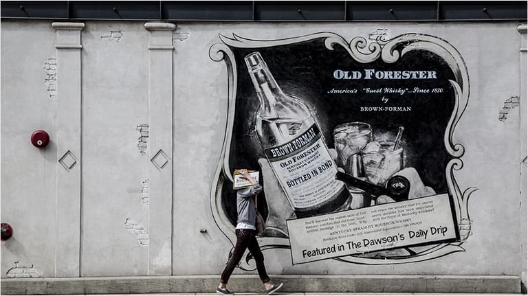 This is an image of a man dressed in black walking in front of a large white billboard/mural. it is advertising Old Forester with an image of a bottle.