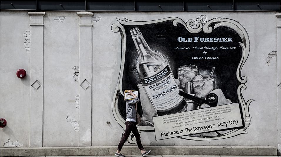 This is an image of a man dressed in black walking in front of a large white billboard/mural. it is advertising Old Forester with an image of a bottle.