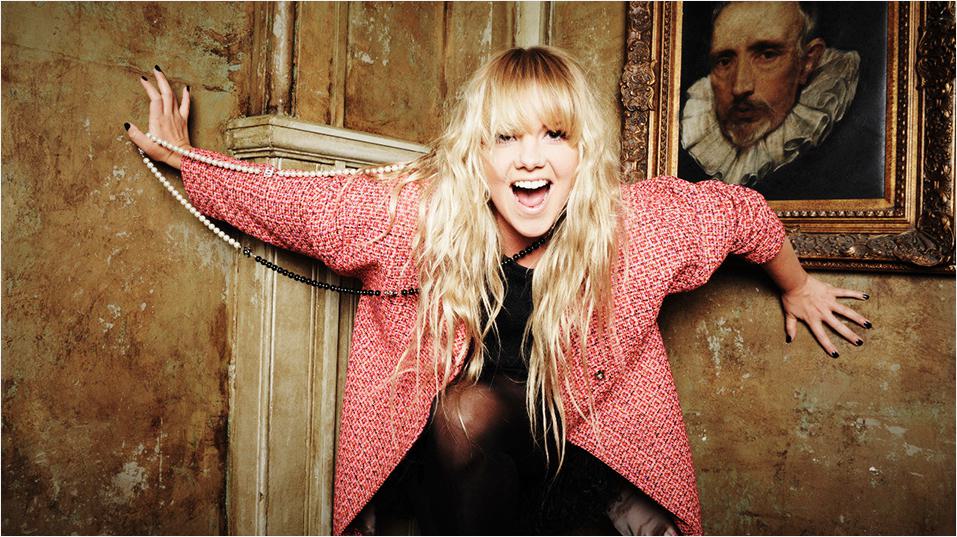 This is a picture of Goldie Rocks, a white woman with long blonde hair and a straight across fringe. She is laughing with her mouth open, with her arms against a wall. Above her is a framed picture of (maybe?) Francis Bacon in a rruff.