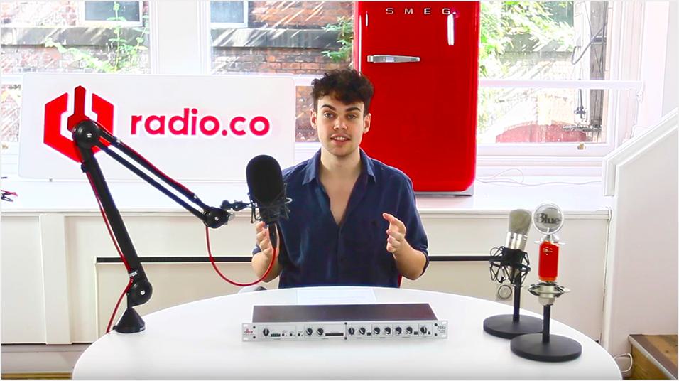 This is a picture of a white man with black hair and a blue shirt sitting in front of a microphone on a boom arm and small mixer. He is pointing at the mic. The back of the image is a red radio.co logo on a white sign.