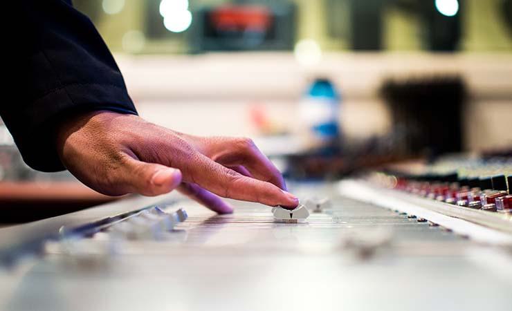 Image shows a hand putting a fader up on a large mixing desk in a radio studio.