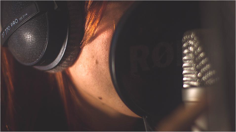This is a picture of a white woman with brown hair on her face. she is wearing black headphones and speaks into a black microphone. We cannot see her facial features.