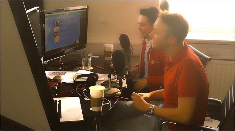 This is a warm, slightly blurred image of two white men sat at a computer screen and mic set up, presumably recording a show together. It is a side on view and one man wears a red shirt.