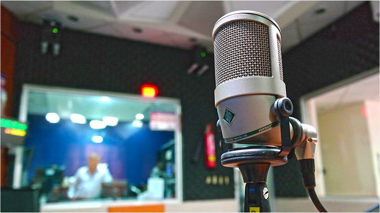This is an image of a silver microphone on a black stand, upright and in the right hand side of the image. In the back is a blue computer screen.