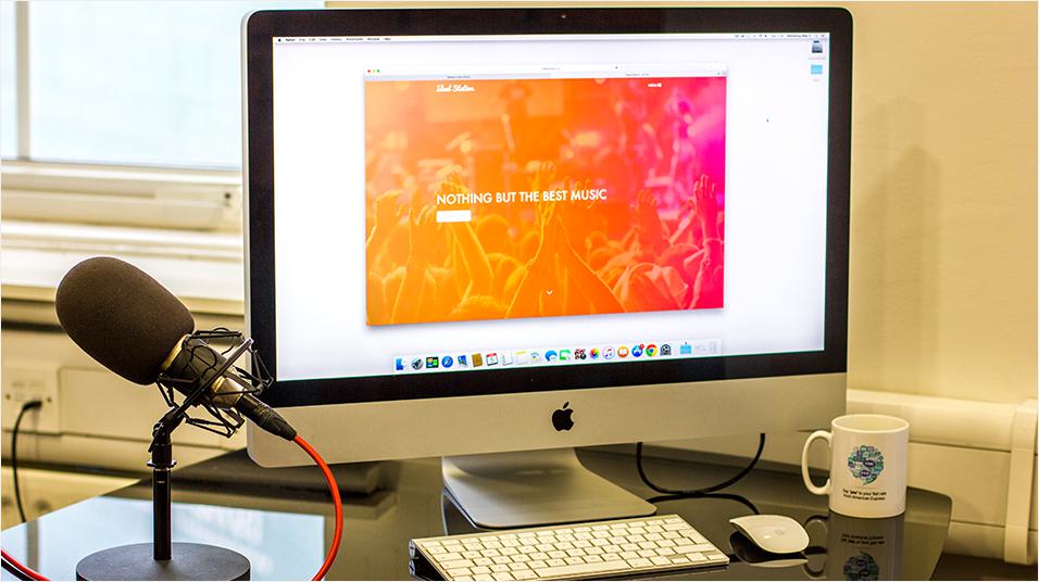 This is a black monitor with a white, pink and orange homepage visible with a taskbar at the bottom. It is a silver Mac. On th eleft there is a black microphone with red wiring.