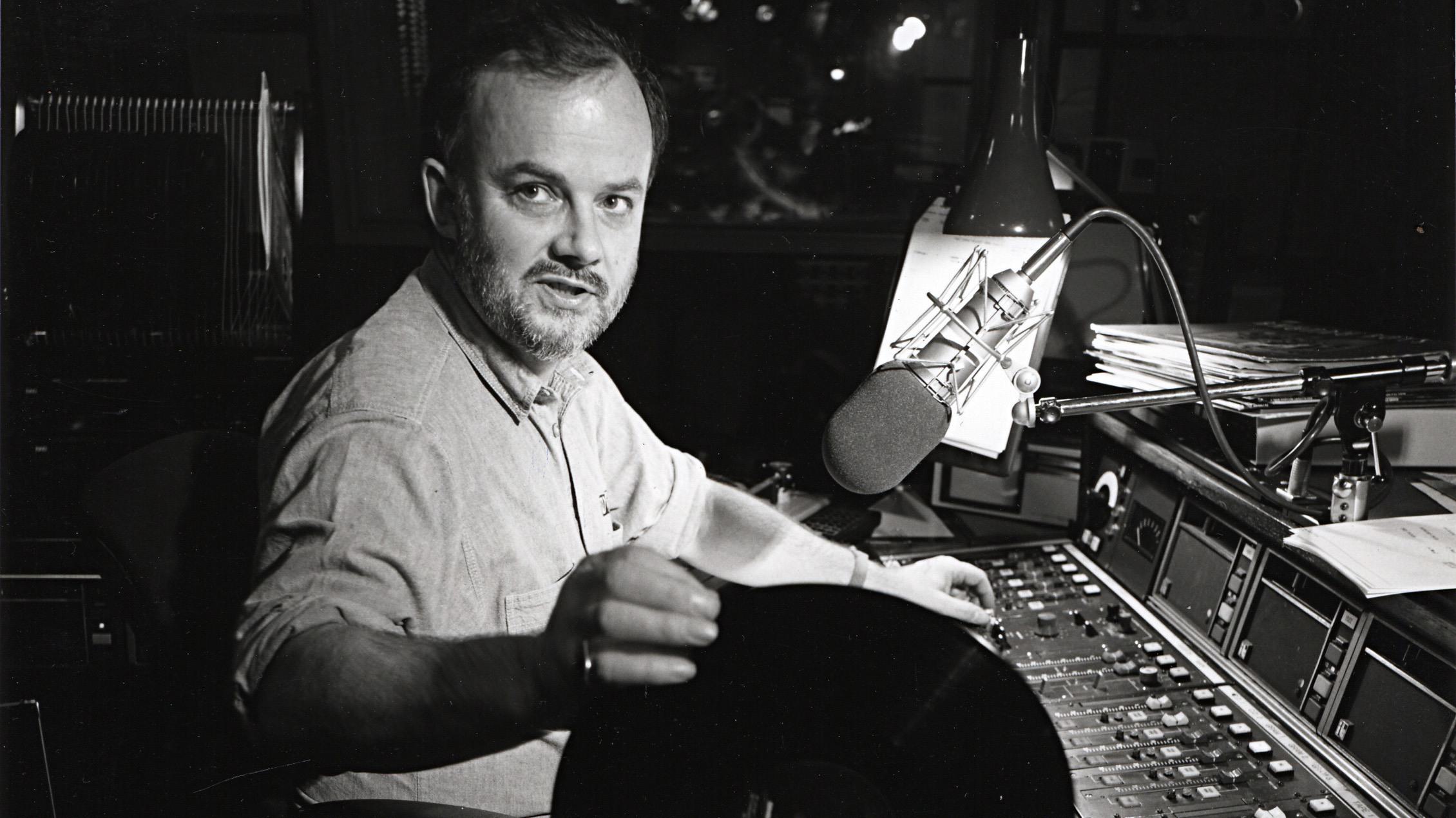 Image is a black and white photo of a young John Peel inside a radio studio.
