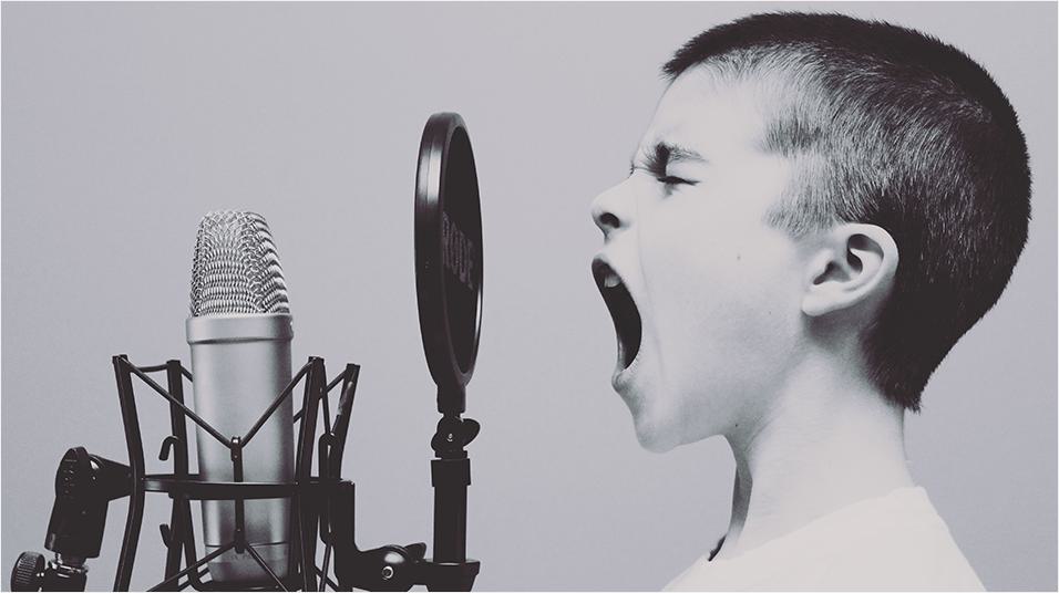 This is a black and white side-on image of a young white boy with a shaved head shouting with his eyes shut into a black mouthguard and microphone.
