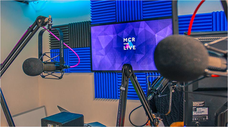 This is an image of the Mcr Live Studio. It has blue soundpanels in the background of the image and a blue monitor reading MCR Live. On the left is a mic on a boom arm, on the right is a mic on the desk. Both have red wires.