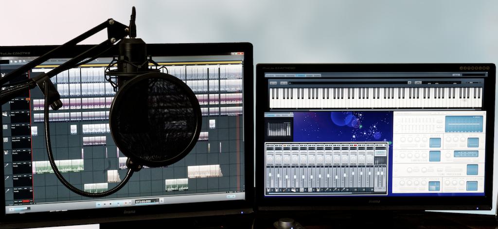 Image shows two computer screens side by side, with different music and audio software on display. In front of the screen is a microphone suspended with a boom arm.