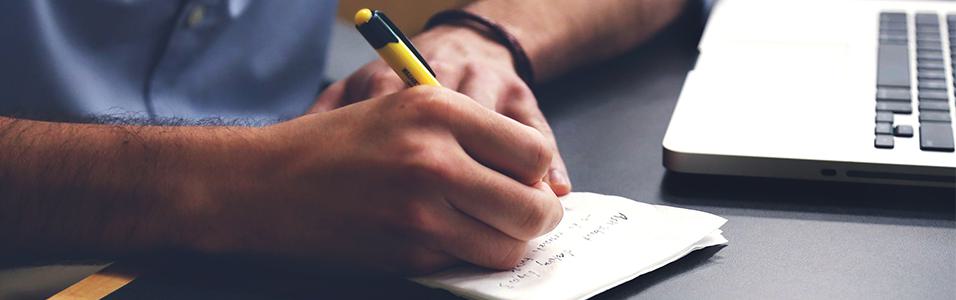 Image shows a close up of a man writing on a notepad, next to a laptop.