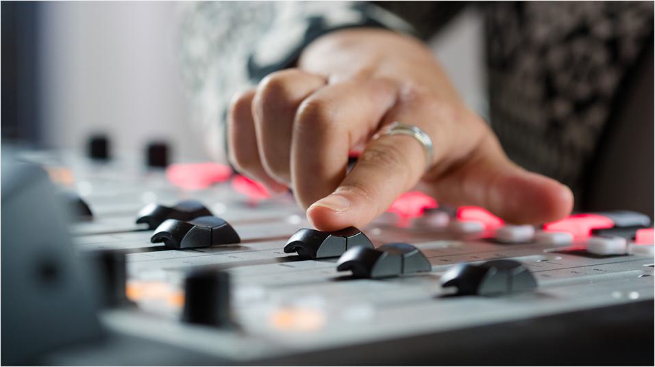 Image is a close-up shot of a hand pushing up a fader on a broadcasting desk.