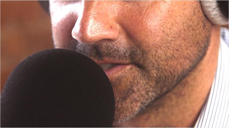 A close up of a white man with a closely shaven beard. He speaks into a rounded black microphone and has headphones on. We can't see anything above his nose.