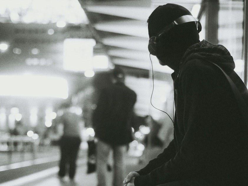Image is a black and white photo of a person listening to headphones at a train station.
