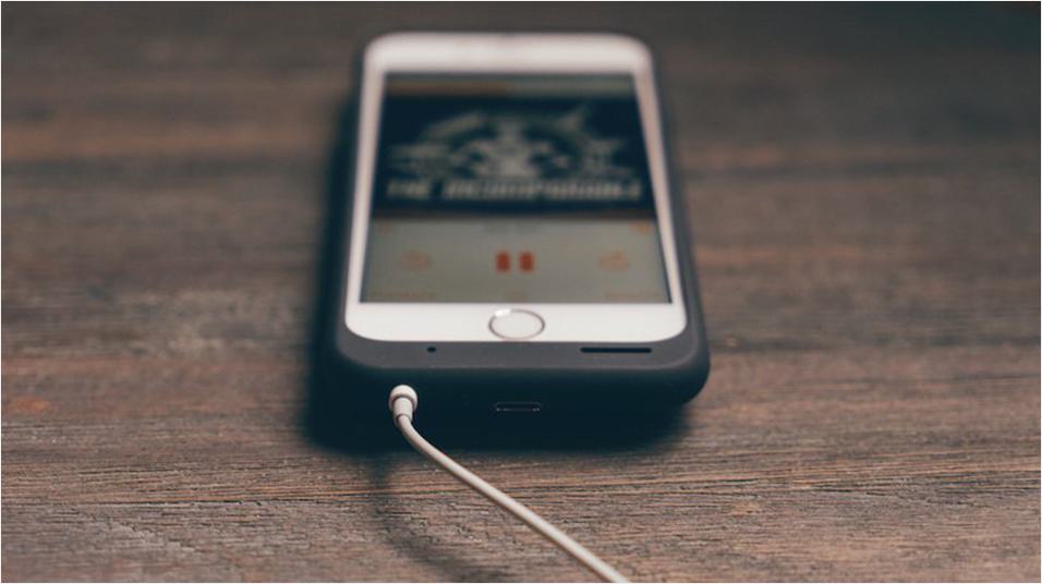 On a dark mahogany backdrop, there is a white iPhone in a case in the middle of the screen.  There is indecipherable album art visible, and a wire, presumably headphones, plugged in.