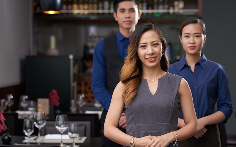 A restaurant host in the foreground, with a male and female waiter behind her.