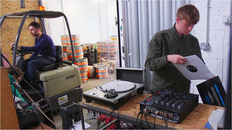 On the right side of the image is a white man with blonde hair putting a record back into its sleeve. he stands in front of a sound mixer and vinyl player. On the left is a white man with dark hair behind the wheel of a moving machine.